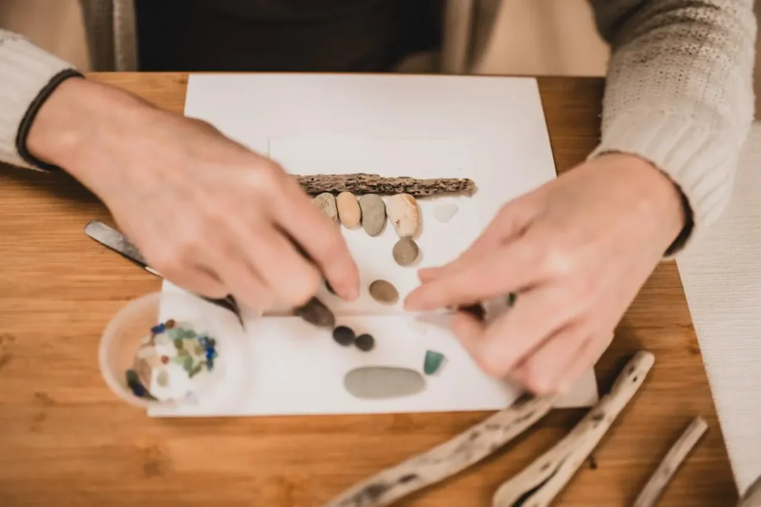 Hands arranging sea glass, pebbles, and driftwood on a canvas
